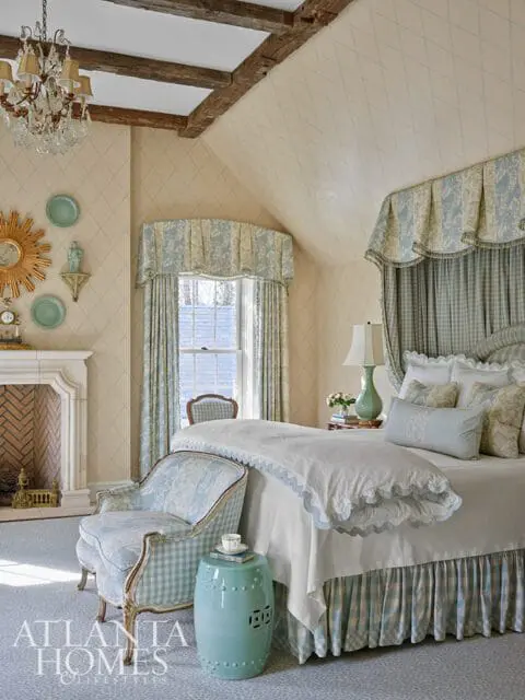 Bedroom with diamond-patterned wallpaper, exposed wooden beams, a crystal chandelier, a fireplace, a patterned armchair, and a bed with blue and white gingham and floral bedding.