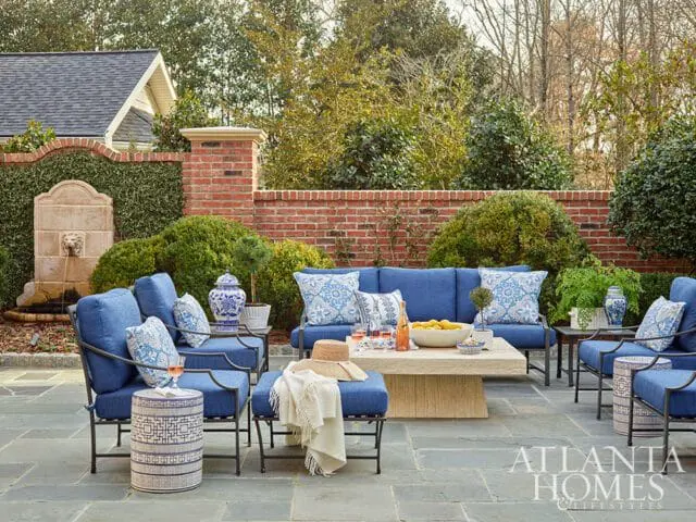 Blue outdoor seating arrangement with patterned pillows and a stone coffee table on a stone patio, with a fountain and brick wall in the background.