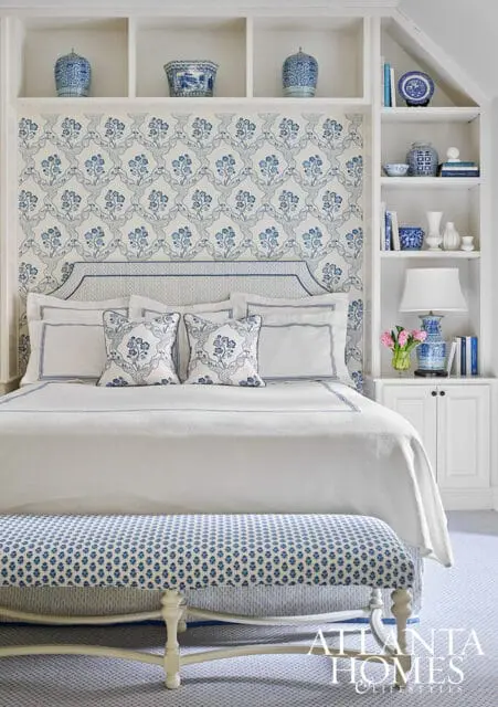 Bedroom with blue and white floral wallpaper, white bedding, and built-in white shelves displaying blue and white decorative vases and books.