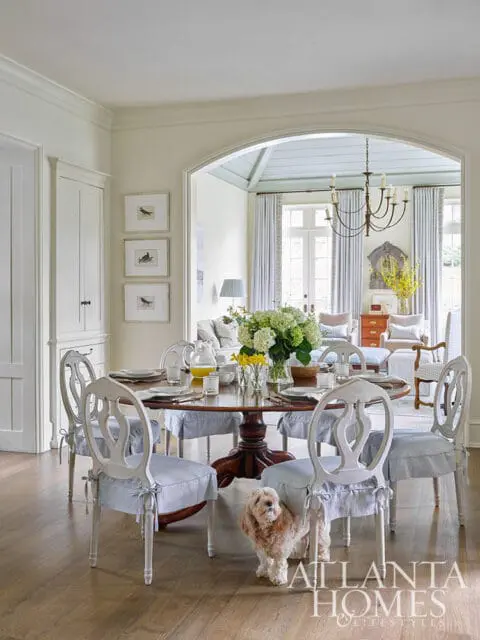 Dining room with round wooden table set for a meal, surrounded by white chairs with blue and white striped cushions. A small dog stands near a chair.