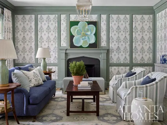 Living room with a blue sofa, striped armchairs, paneled walls with floral wallpaper, a fireplace with a floral painting, and a patterned rug.