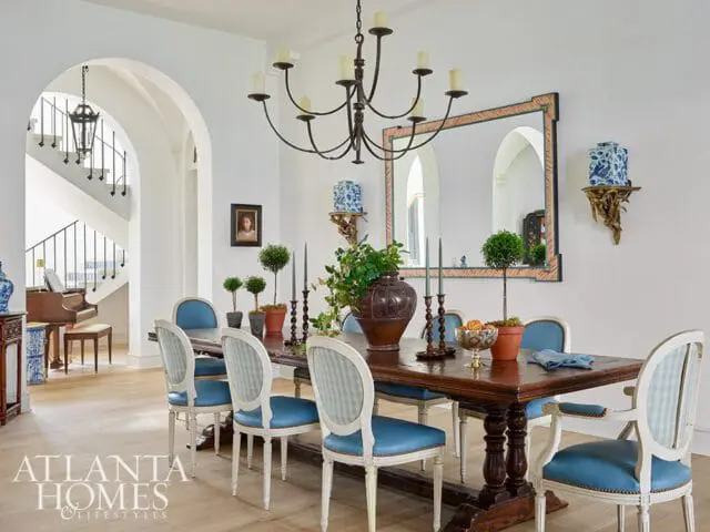 Dining room with a dark wood trestle table set with blue upholstered chairs, a large ornate mirror, and a grand staircase with a piano in the background.