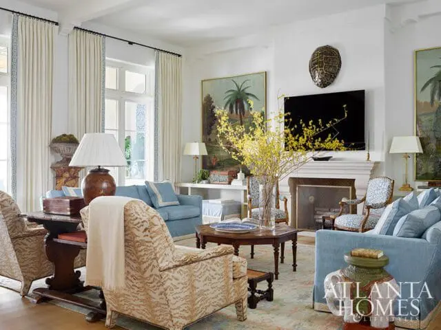 Living room with light blue sofas, patterned armchairs, a fireplace with a turtle shell above the TV, and tall windows with white curtains featuring blue trim.