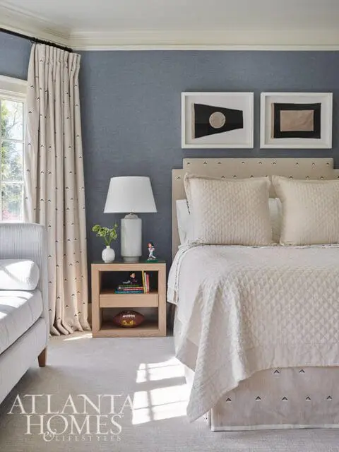 Bedroom with blue textured wallpaper, a bed with a neutral upholstered headboard and quilted bedding, and a wooden nightstand with a lamp and decorative items.