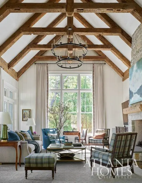 Living room with vaulted ceiling featuring exposed wooden beams, a large window overlooking trees, a stone fireplace, and plaid upholstered furniture.