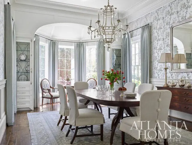 Elegant dining room with a dark wood oval table, upholstered chairs, a crystal chandelier, and a mirrored wall with decorative lamps and a dresser.