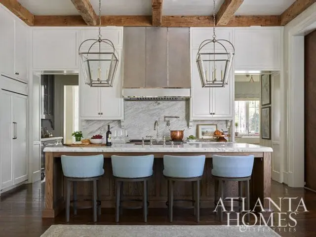 Kitchen island with light blue upholstered bar stools, marble backsplash, and stainless steel range hood. White cabinetry and exposed wooden beams overhead.