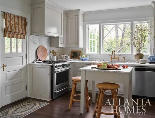 White kitchen with marble countertops, stainless steel range, and a small island with woven stools. A window with diamond panes overlooks greenery.