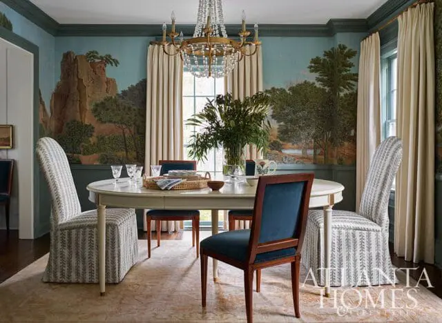 Dining room with a large oval table set for four, upholstered chairs with patterned skirts, and a scenic mural wallpaper depicting a landscape with cliffs and trees.