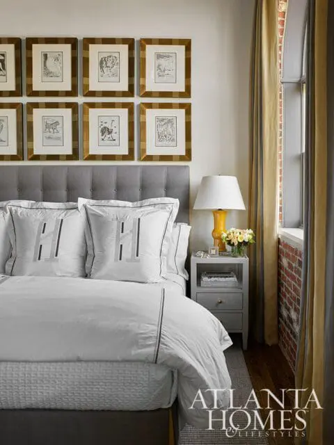Bedroom with a tufted gray headboard, white bedding with monogrammed pillows, and a gallery wall of framed botanical prints. A bedside table holds a lamp and flowers.