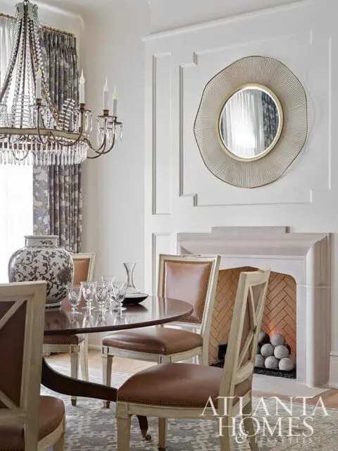 Dining room with a round wooden table, brown leather chairs, a crystal chandelier, a fireplace with a decorative mirror above, and a large patterned vase.