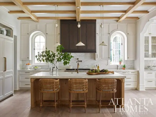 White kitchen with wood-beamed ceiling, marble countertops, a dark wood range hood, and woven bar stools at a central island.
