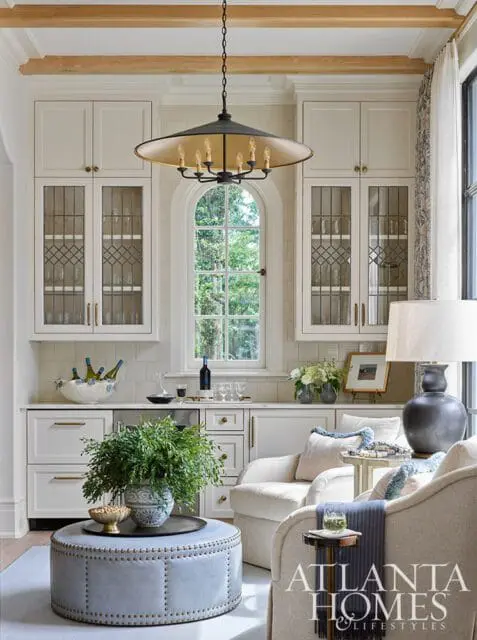 Creamy white kitchen with arched window, glass-front cabinetry, and a large black chandelier. A blue tufted ottoman sits in the foreground with two chairs.