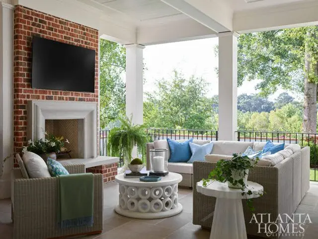 Covered patio with brick fireplace, TV, sectional sofa with blue pillows, wicker armchairs, and round white coffee table with decorative base.
