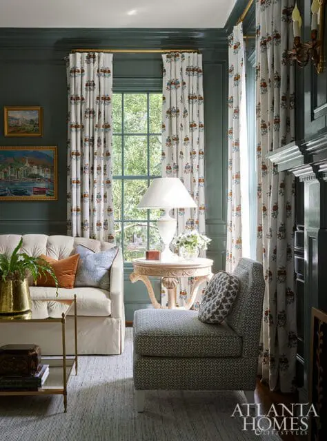 Living room with dark green walls, cream sofa with orange and blue pillows, patterned curtains, and a small armchair with patterned pillow.