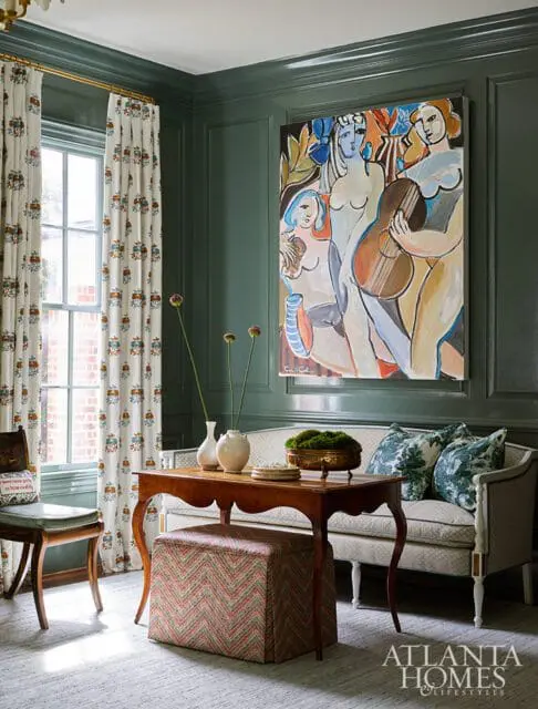 Dark green paneled walls with a large abstract painting of figures and a guitar above a sofa. A wooden table with vases and an ottoman sits in front of the sofa.