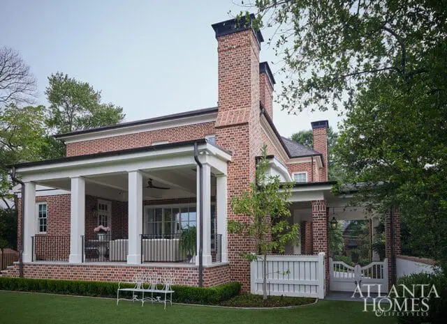 Red brick house with white columns supporting a covered porch, black metal railings, and two chimneys. A white picket fence and gate lead to the entrance.