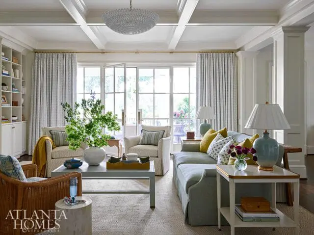 Living room with coffered ceiling, large windows, a light blue sectional sofa with yellow and patterned pillows, and a wicker armchair.