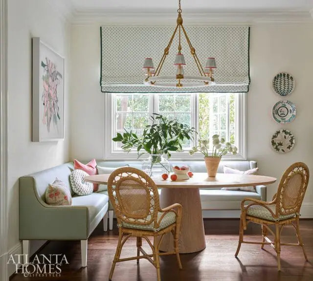 Light blue banquette seating with decorative pillows surrounds an oval wood dining table with two rattan chairs. A patterned roman shade covers a large window.
