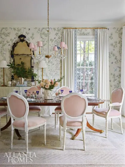 Dining room with a dark wood table set for a meal, surrounded by white and pink upholstered chairs. A floral wallpaper and ornate mirror adorn the walls, with a crystal chandelier overhead.