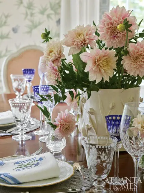 Dining table set with pink dahlias in a white vase, crystal glassware, blue-accented wine glasses, and embroidered napkins.