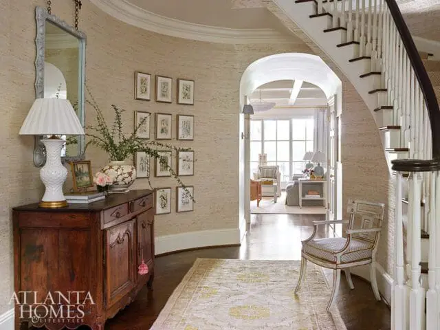 Foyer with grasscloth wallpaper, a dark wood antique console table with a white lamp and floral arrangement, a gallery wall of botanical prints, and a curved staircase with white balusters.