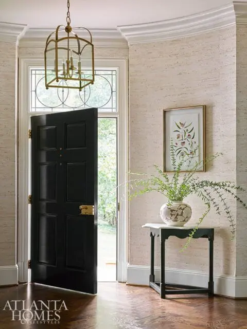 Entryway with a black paneled door, brass lantern, textured wallpaper, and a small console table with a vase of greenery.