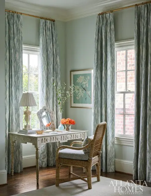 Light green room with a vintage desk, wicker chair, and patterned blue curtains. A framed bird print hangs on the wall.