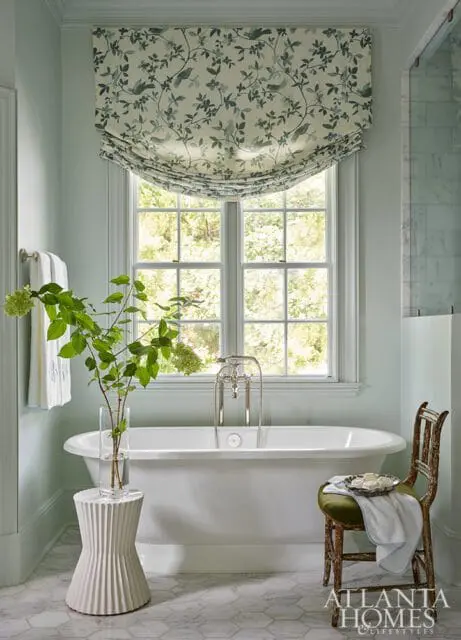 Light blue bathroom with a freestanding white bathtub, a floral Roman shade window treatment, and a vintage wooden chair with an olive green seat.