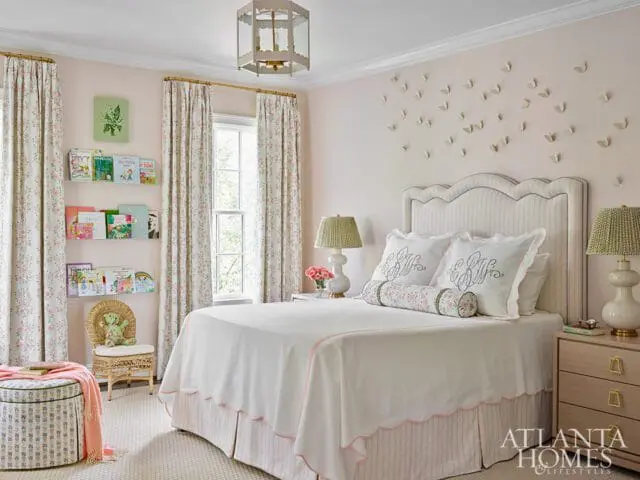 Children's bedroom with pink walls, a scalloped upholstered headboard, white bedding with pink trim, and floral curtains. Books are displayed on clear shelves.