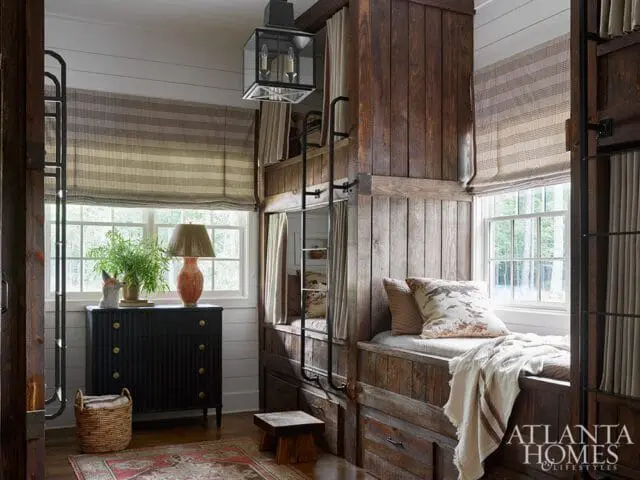 Bedroom with built-in wooden bunk beds, dark dresser with a lamp and plant, and striped Roman shades over windows.