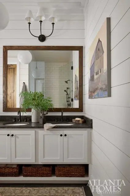White shiplap bathroom with a double vanity featuring white cabinets, dark granite countertops, black faucets, and woven storage baskets below. A large wood-framed mirror reflects a subway tile shower.