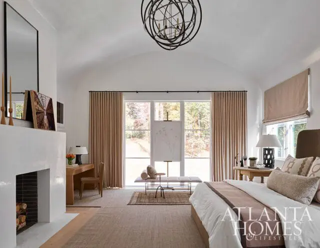 Bedroom with vaulted ceiling, white plaster fireplace, wood desk, and bed with neutral bedding. Large windows overlook a wooded area.