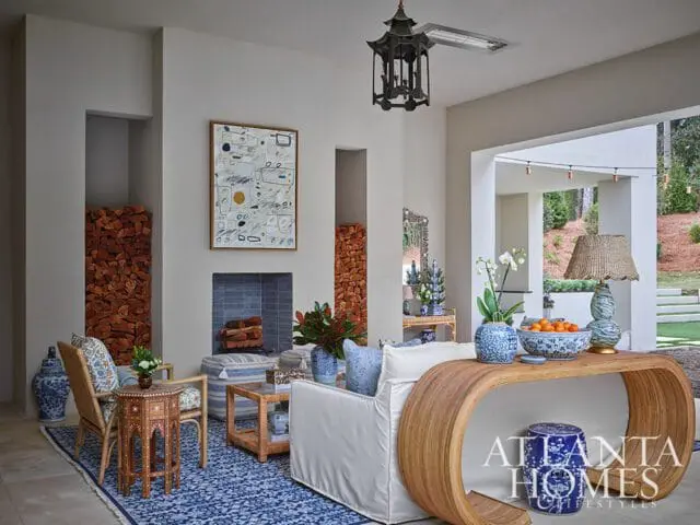 Living room with a white sofa, rattan console table, blue and white patterned rug, wicker chairs, and a fireplace with stacked logs.