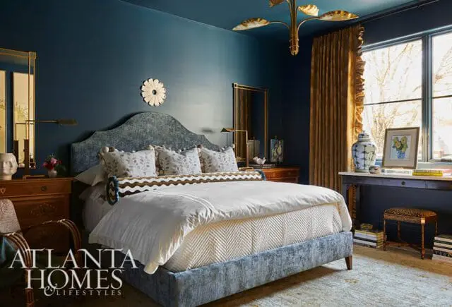 Bedroom with dark blue walls, a blue upholstered bed with patterned pillows, white bedding, and gold curtains. A wooden dresser and a desk with a ginger jar and artwork are visible.