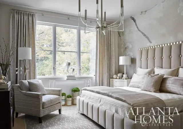 Neutral bedroom with tufted headboard, plush bedding, patterned armchair, and large window overlooking trees. Atlanta Homes & Lifestyles logo visible.