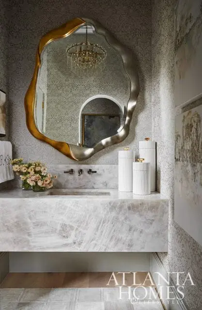 Powder room with a marble vanity, ornate gold and silver mirror reflecting a crystal chandelier, and textured wallpaper.