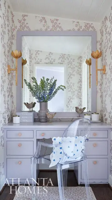 Lavender vanity with pink knobs, clear acrylic chair with blue heart pillow, floral wallpaper, and gold sconces.