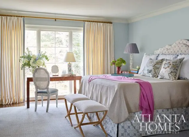 Light blue bedroom with a queen-size bed, patterned headboard, and a desk with a cane-back chair. Cream curtains frame a large window.