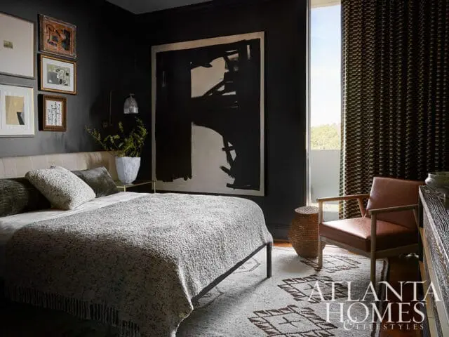Dark bedroom with a large abstract painting, a textured bed with grey bedding, a leather armchair, and a patterned rug with the Atlanta Homes & Lifestyles logo.