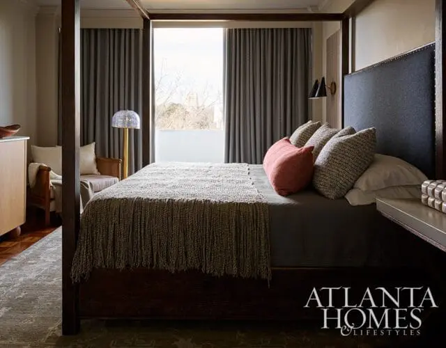 Bedroom with a dark wood four-poster bed, upholstered headboard, textured throw blanket, and patterned pillows. Dark gray curtains frame a large window.