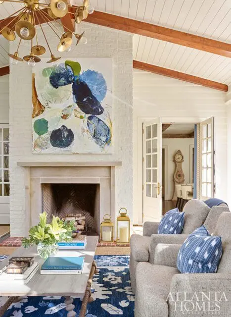 Living room with white brick fireplace, abstract art, vaulted ceiling with wood beams, and upholstered chairs with blue patterned pillows.