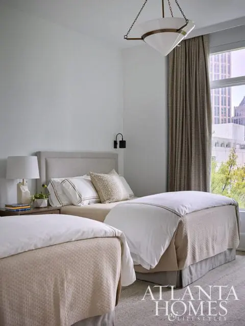 Bedroom with two twin beds, neutral bedding, upholstered headboards, a modern chandelier, and patterned curtains revealing a city view.