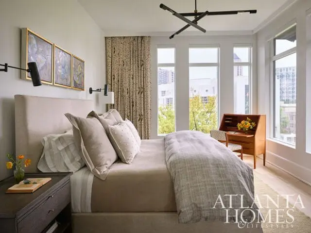 Bedroom with a neutral upholstered bed, patterned curtains, three framed artworks, a wooden secretary desk, and large windows overlooking a city skyline.