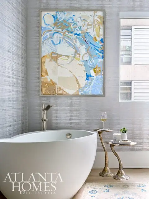 Modern bathroom with a freestanding white soaking tub, abstract art on textured silver wallpaper, and a glass of white wine on a sculptural side table.