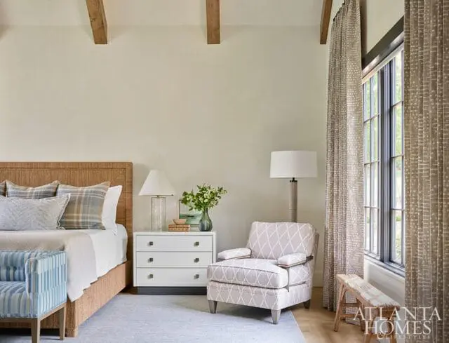 Bedroom with a woven headboard, plaid pillows, a white dresser, and a patterned armchair. Exposed wooden beams accent the ceiling.