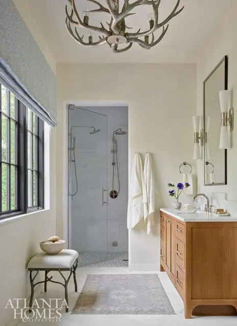 Cream bathroom with a glass-enclosed shower, wooden vanity with marble countertop, antler chandelier, and a window with blue patterned Roman shades.