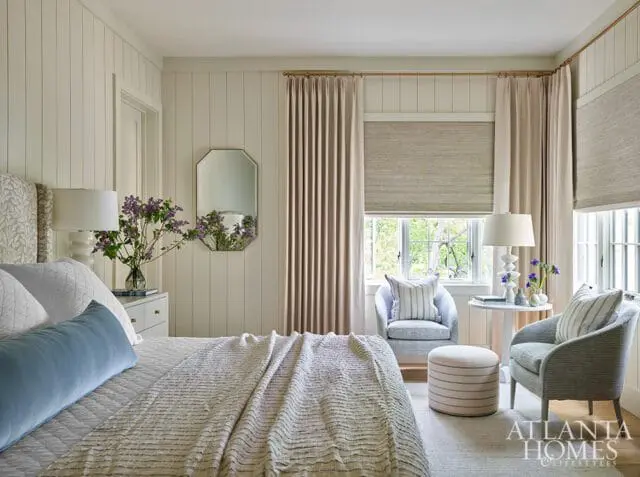 Bedroom with shiplap walls, a bed with a blue bolster pillow and striped throw, two armchairs, and a small round table with lamps and flowers.