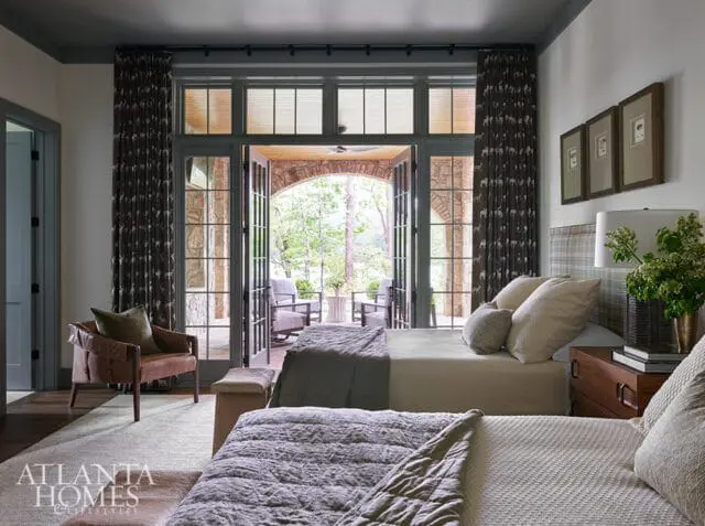 Bedroom with two beds, plaid headboard, leather armchair, and French doors opening to a stone patio with trees beyond.