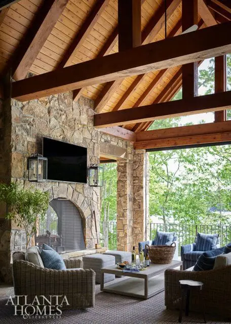 Covered patio with stone fireplace, television, wicker furniture, and a view of trees and water.